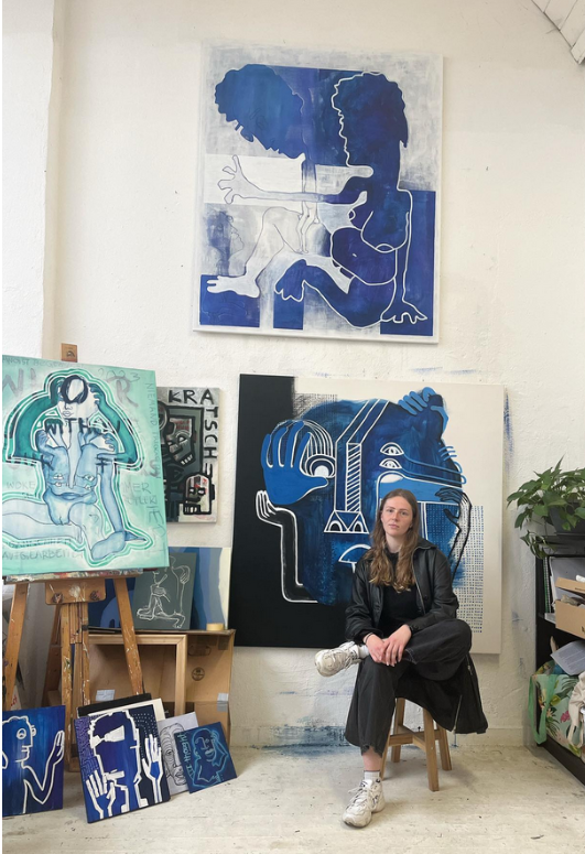 Künstlerin Elisa Zelmerin ihrem Atelier, umgeben von mehreren großformatigen, blau dominierten Gemälden in abstrakter Figuration. Sie sitzt entspannt vor einem ihrer Werke, während weitere Leinwände und Skizzen den kreativen Raum füllen.